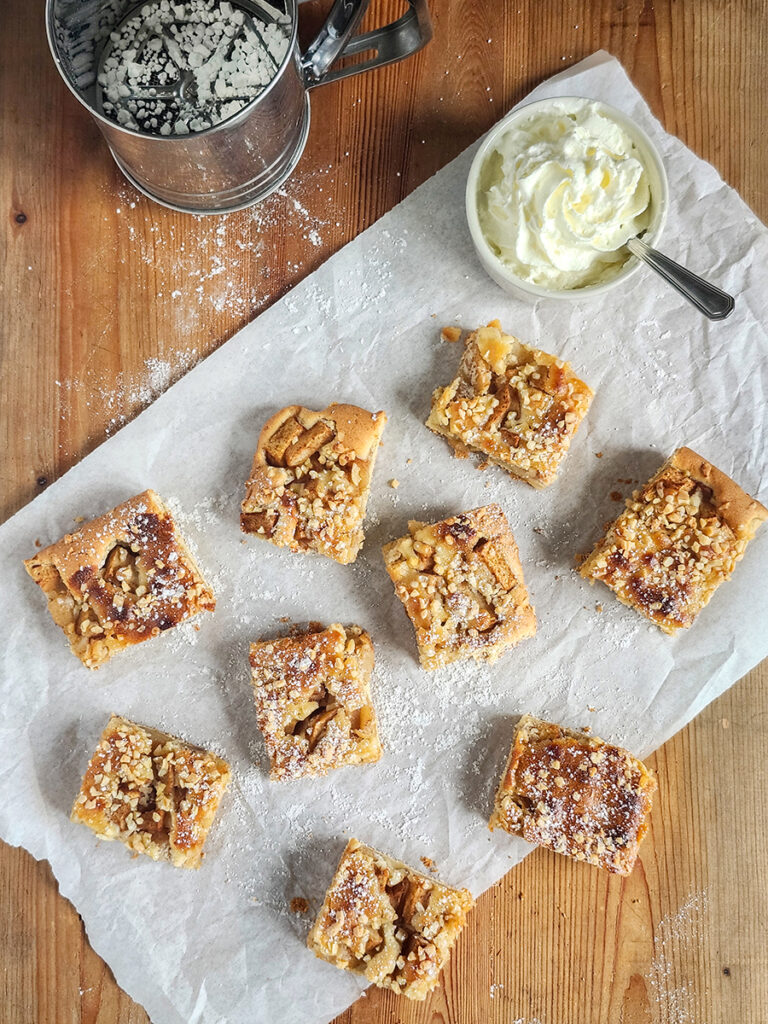 Mehrere Stücke Apfelkuchen vom Blech mit Marzipan, Puderzucker und Sahne auf Backpapier