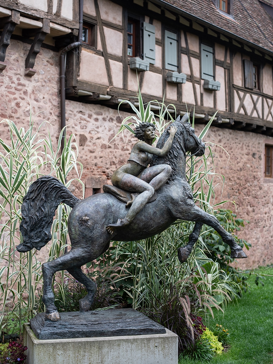 Bronze-Statue eines Mädchens auf einem Pferd vor Fachwerkfassade in Riquewihr