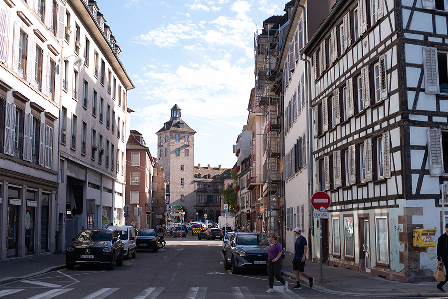 Straßenszene in Straßburg mit Autos, Fachwerkhäusern und einem historischen Turm im Hintergrund