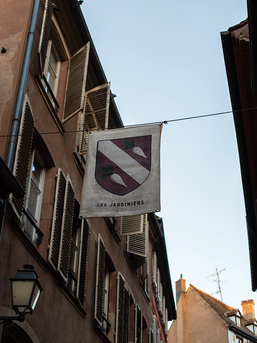 Banner mit Wappen und Aufschrift „Les Jardiniers“ hängt in einer Gasse in Straßburg