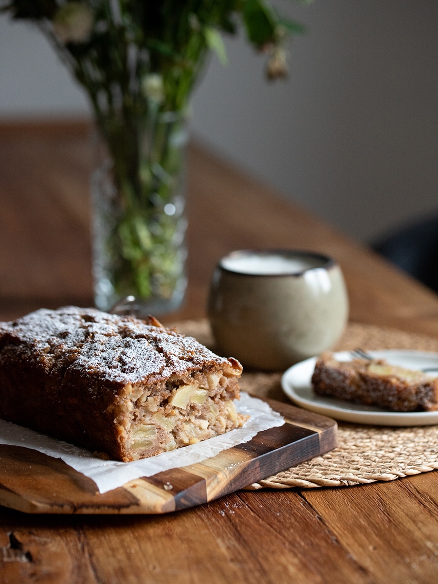 Kleiner Apfel-Mandel-Kuchen mit Puderzucker auf einem Holzbrett, daneben eine Vase mit Blumen – gemütliche Kaffee-Atmosphäre