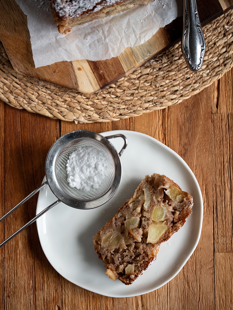 Ein Stück Apfel-Mandel-Kuchen auf einem Teller mit Gabel, daneben die kleine Kastenform auf einem Holztisch.