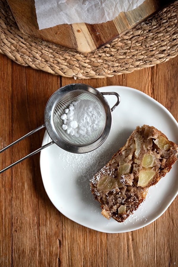 Ein Stück Apfel-Mandel-Kuchen mit Puderzucker auf einem weißen Teller, daneben ein Teesieb und Besteck auf einem rustikalen Holztisch.