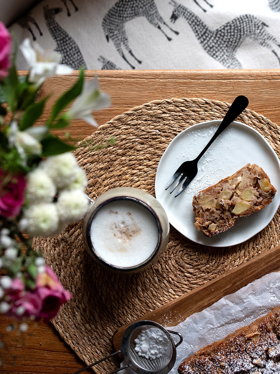 Ein Stück Apfel-Mandel-Kuchen mit Puderzucker auf einem Teller, daneben eine Tasse Kaffee und Besteck auf einem rustikalen Holztisch.