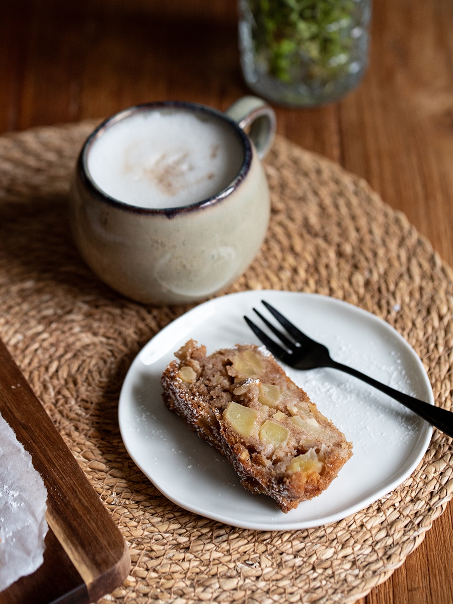 Ein Stück Apfel-Mandel-Kuchen auf einem Teller mit Gabel, daneben eine Tasse Kaffee auf einem Holztisch.