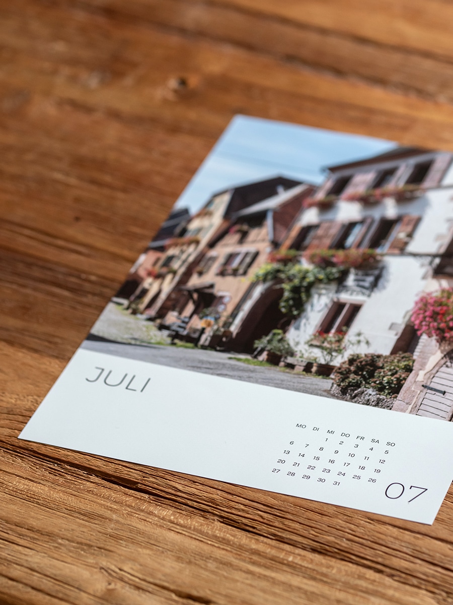 Aufgeschlagener Elsass-Kalender 2026, der ein Dorfmotiv mit Fachwerkhäusern und Blumen zeigt.