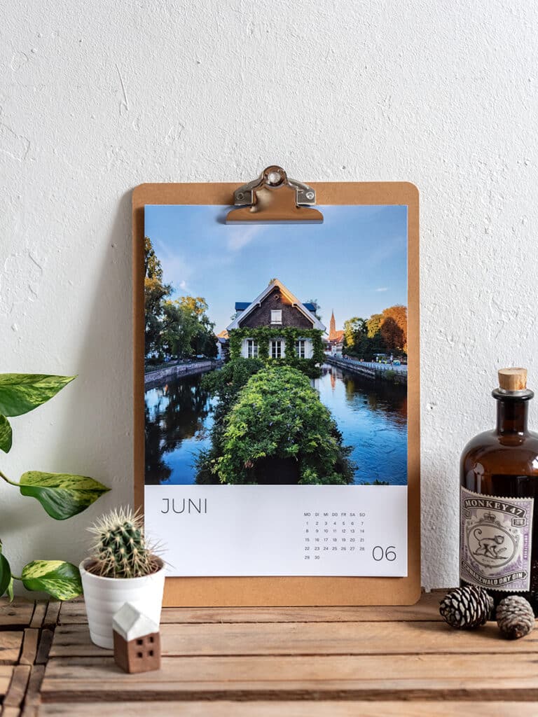 Wandkalender ‚Elsass 2026‘ auf einem Holzregal, mit einem Deckblattmotiv aus Berglandschaft, Wasser und Fachwerkhäusern.