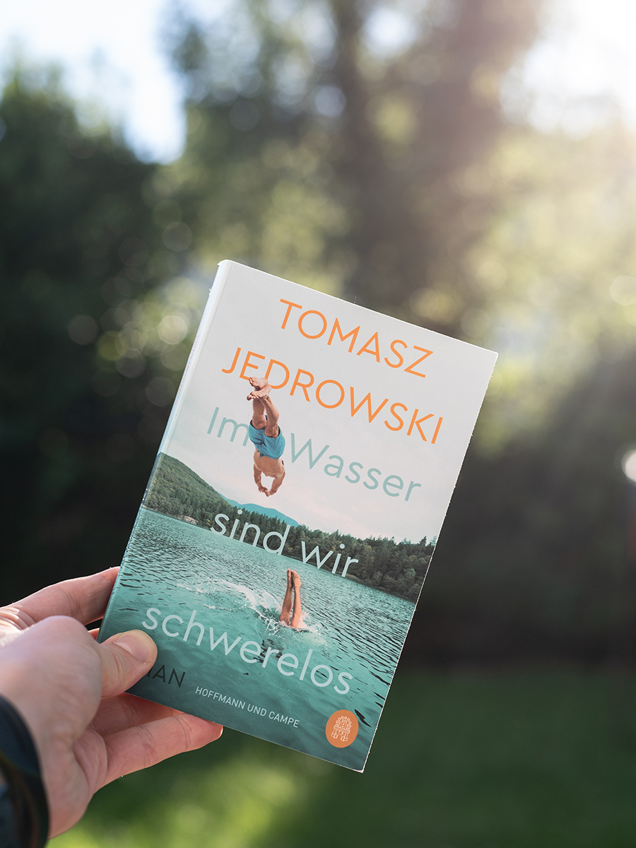Hand hält das Buch „Im Wasser sind wir schwerelos“ von Tomasz Jedrowski vor unscharfem grünem Hintergrund