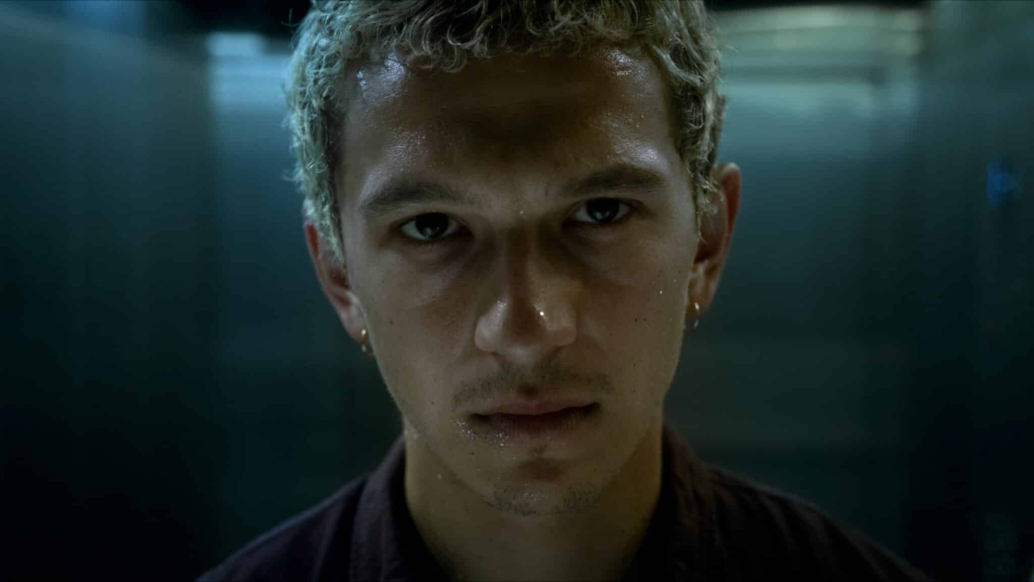 Filip blickt mit ernstem Gesichtsausdruck in die Kamera. Die Szene ist in kühlen Blau- und Grautönen gehalten und stammt aus der HBO-Max-Serie „Proud“.