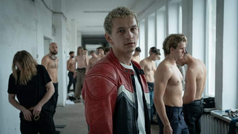 Filip steht in einem Umkleideraum. Im Hintergrund sind weitere Models zu sehen, die sich auf ein Casting oder Shooting vorbereiten.